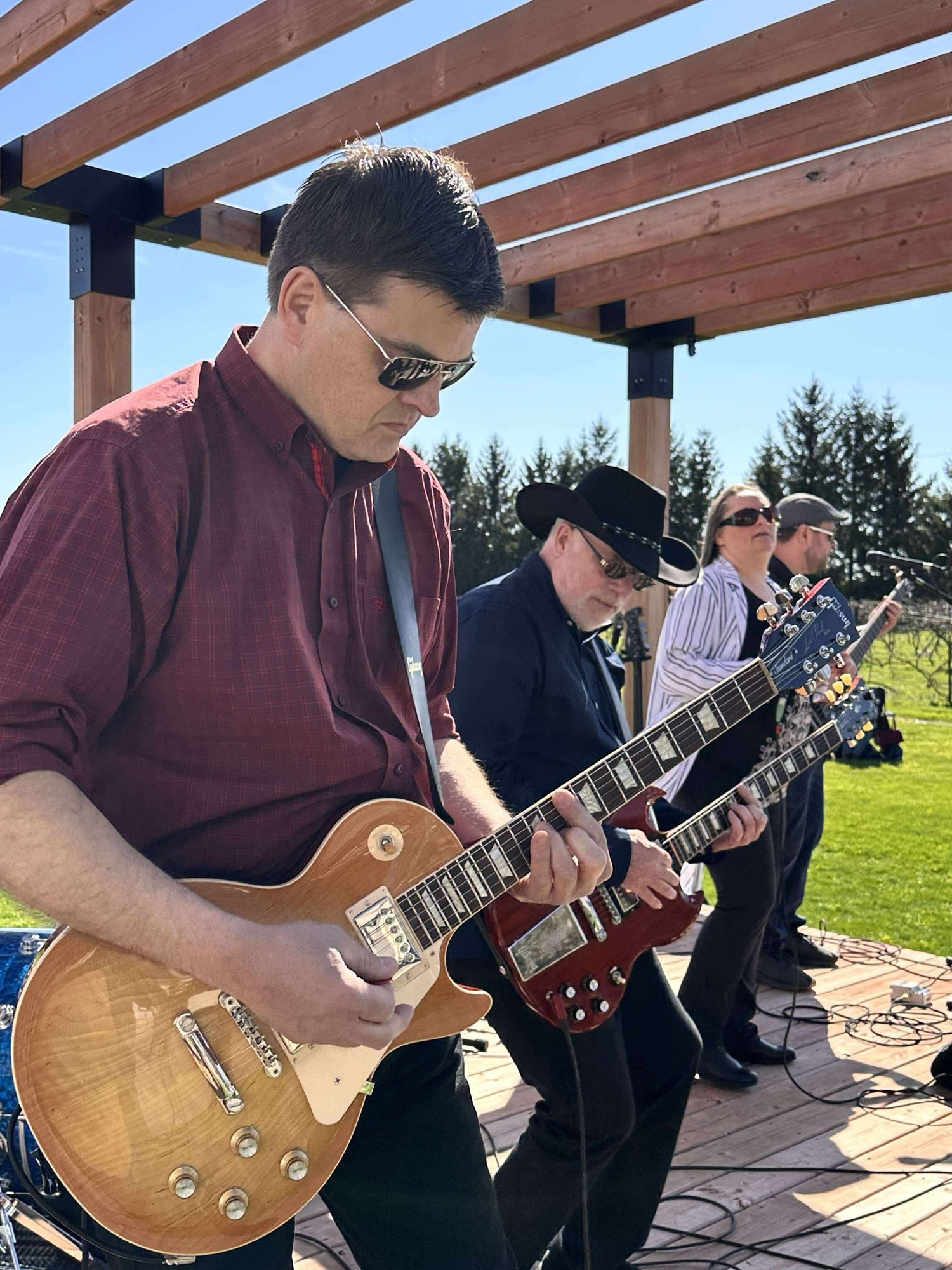 Ross Burt with a Les Paul, Brent Rougeau in a cowboy hat with a red Gibson SG, performing under a vineyard pergola on a sunny afternoon